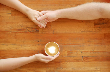 Young couple in love holding hands over red cup of coffee with heart shaped foam latte art on vintage grunged scratched table. Man & woman body language. Background, top view, close up, copy space.