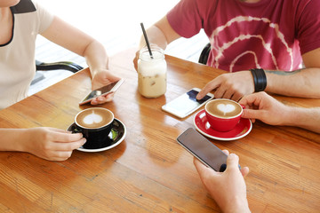 Group of friends drinking cappuccino at coffee shop, staring at smartphones, checking likes & messages. Addiction to social media. Young men & women holding cell phones & drinks. Background, top view.