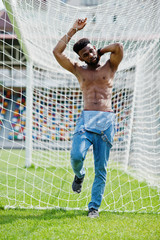 Handsome sexy african american muscular bare torso man at jeans overalls posed at green grass agasinst football gates of stadium field. Fashionable black man portrait.