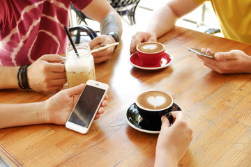 Group of friends drinking cappuccino at coffee shop, staring at smartphones, checking likes & messages. Addiction to social media. Young men & women holding cell phones & drinks. Background, top view.