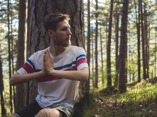 young bearded man meditating sitting on ground under the tree in pine forest on summer day