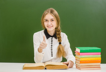 Smiling teen girl with books on the background of a school board showing thumbs up