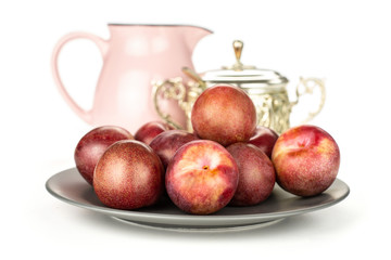 Lot of whole fresh pluot interspecific plums variety on a ceramic grey plate with a pink jar and sugar bowl still life isolated on white