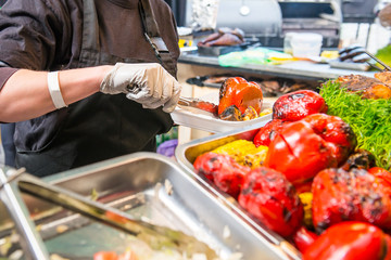 Close up female seller hands holding grilled bell peper in the takeaway food container. A buffet on a holiday on the street food culinary market, festival, event. Selective focus. copy space.
