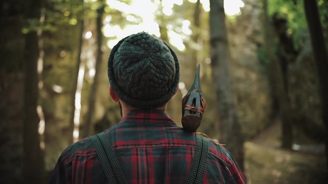 Walking bearded lumberjack with axe. Man in a cap walks through the woods in search of the tree. View behind the back