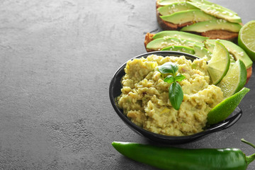 Bowl with tasty guacamole and toast with sliced avocado on grey textured background