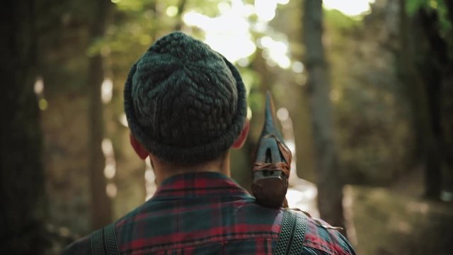 Walking bearded lumberjack with axe. Man in a cap walks through the woods in search of the tree. View behind the back