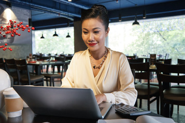 Fototapeta premium Happy Asian businesswoman in white blouse sitting at wooden table in restaurant and using laptop on blurred background 