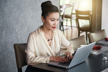 Smiling attractive Asian businesswoman typing on laptop keyboard sitting comfortably on chair at dark wooden table in restaurant on blurred background 