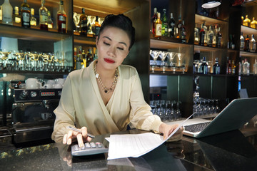 Beautiful Asian woman bartender concentrating on using calculator doing business accounts and...