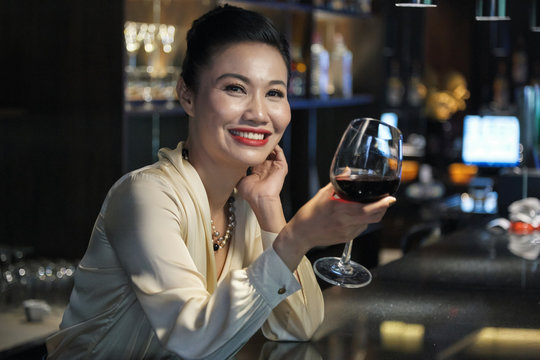 Side view of beautiful cheerful Asian businesswoman in light blouse relaxing with glass of wine in bar and looking at camera smiling on blurred background