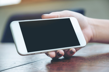 Mockup image of a woman holding and showing white mobile phone with blank black screen for watching on wooden table