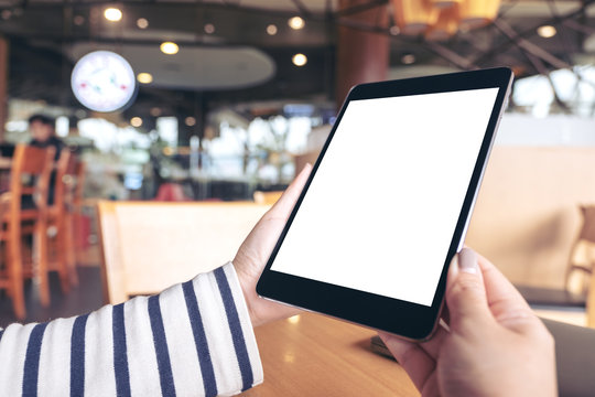 Mockup image of hands holding black tablet pc with blank white desktop screen on wooden table in cafe