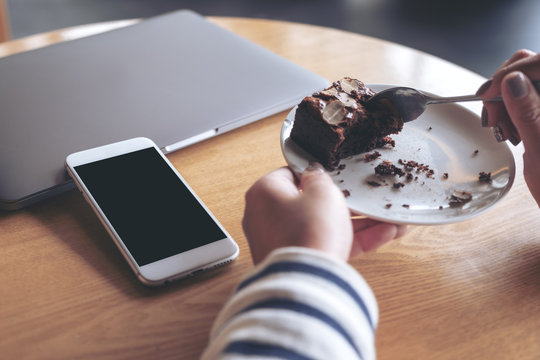 Mockup image of a white mobile phone with blank black desktop screen next to laptop with a woman eating brownie cake