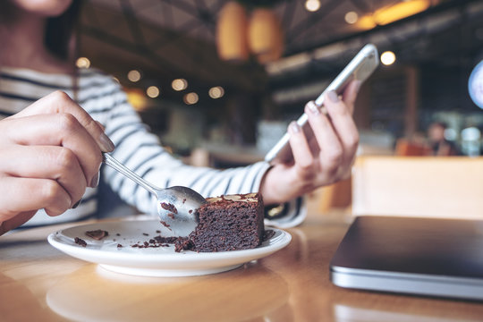 Closeup image of a woman holding , using and looking at smart phone while eating brownie cake in cafe