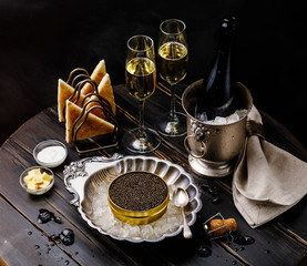 Black caviar on ice in silver bowl, fresh bread toast and champagne in ice bucket on dark background