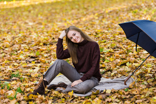 Pretty teenager girl sits on yellow autumn leaves on the ground in park and drinks tea