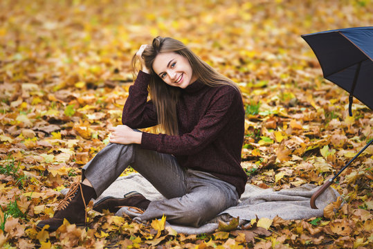 Pretty teenager girl sits on yellow autumn leaves on the ground in park and drinks tea