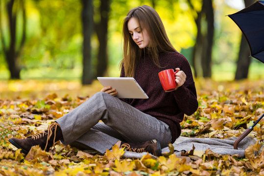 Pretty teenager girl  is  usind gudget sitting  on yellow autumn leaves on the ground in park and drinks tea