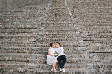 Fototapeta na wymiar Beautiful newlyweds are sitting on old steps and embrace. A stylish bridegroom hugs a cute bride against the backdrop of a staircase.