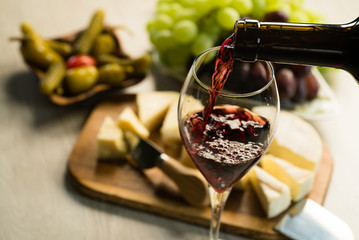 Obraz premium Pouring red wine into the glass in the background a table set with various types of cheese