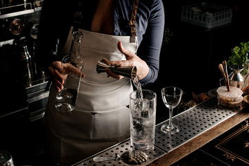Female bartender holding a bottle and pouring an alcohol into the glass with ice
