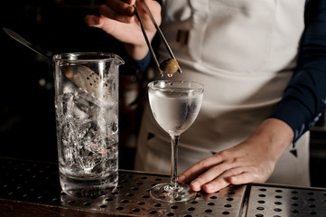 Female bartender adding to the cocktail one olive with tweezers