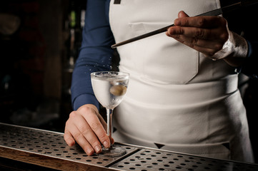 Bartender girl adding into the transparent cocktail one olive with tweezers