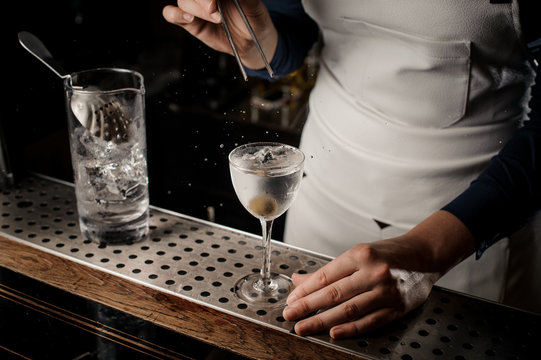 Female bartender throwing into the transparent cocktail one olive with tweezers