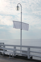 Mock-up of a white flagpole on a pier near the calm sea and cloudy sky. Banner for your design