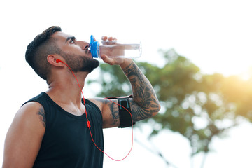 From below side view of tattooed sportsman in headphones and armband drinking water from bottle on...