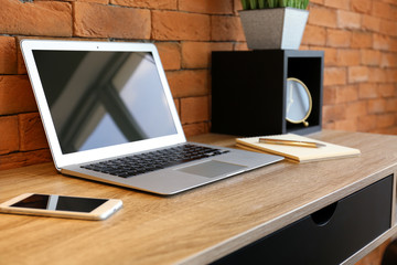 Modern laptop with mobile phone on wooden table near brick wall in office