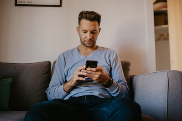 Young handsome man sitting at home on sofa and using mobile phone. Men holding smartphone hands and typing text message