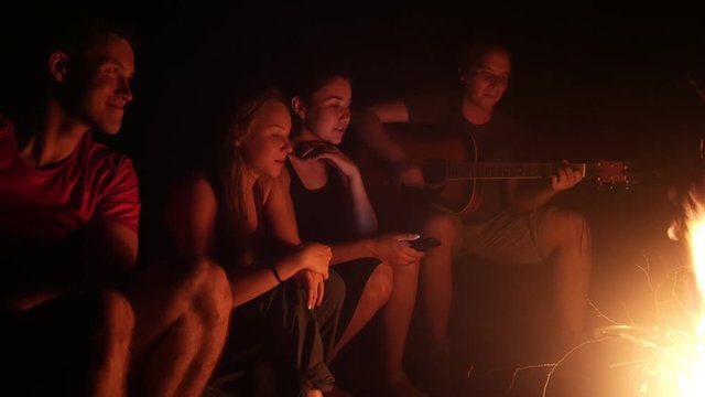 Young friends sitting near the campfire and singing songs with a guitar at night