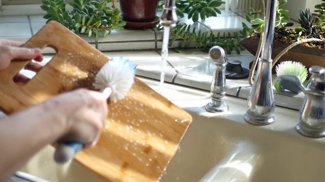 Chef washing wooden cutting board with soap and water in kitchen sink.