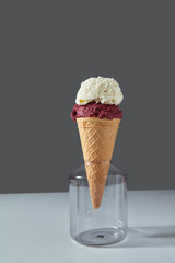 A balls of berry and vanilla ice cream in a waffle cone stands in a glass jar on a gray background on the table.