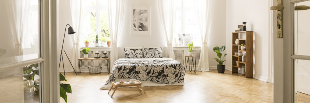 Glass door entrance into a beautiful, bright bedroom interior with breakfast tray on a wooden floor and black floral pattern sheets on a comfy bed