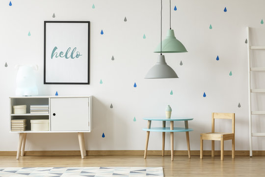 Real photo of a pastel blue and mint kid room interior with a poster above a white cupboard standing next to a round, wooden table