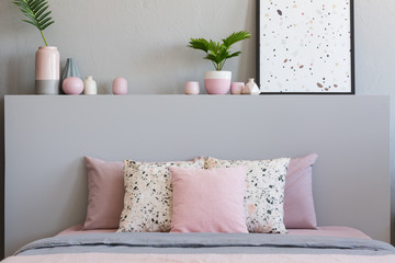 Pink and patterned pillows on bed with headboard in grey bedroom interior with poster. Real photo