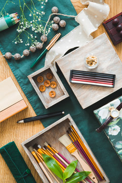Boxes with brushes and buttons placed on wooden table with green cloth, fresh flowers and pencil crayons in photo with high angle