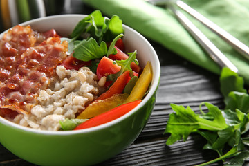 Bowl with delicious oatmeal, bacon, herbs and vegetables on wooden table, closeup