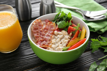 Bowl with delicious oatmeal, bacon, herbs and vegetables on wooden table