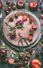 Wooden plate with seasonal fruits, berries, flowers and scissors on rustic kitchen table background, top view. Country style