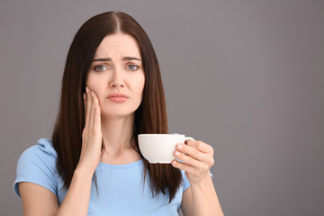 Young woman with sensitive teeth and cup of hot coffee on grey background