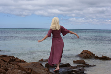 full length portrait of blonde girl wearing purple dress, walking along a beach.