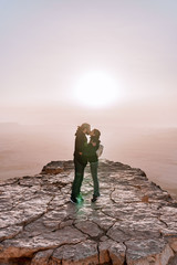 Fototapeta premium Alone young couple of man and women with backpack in israel negev desert admires the view of sunrise. Young pair stands on the edge of the cliff of makhtesh ramon park.