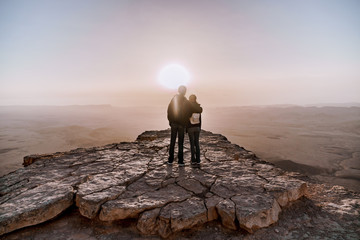 Alone young couple of man and women with backpack in israel negev desert admires the view of sunrise. Young pair stands on the edge of the cliff of makhtesh ramon park. - obrazy, fototapety, plakaty