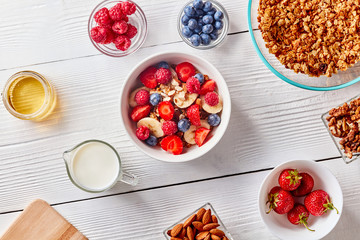 Ingredirnts for preparing healthy organic breakfast - granola, berries honey, milk, banana in white bowl on white table. Flat lay.