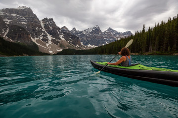 Adventurous woman kayaking in Moraine Lake during a striking cloudy sunset. Taken in Banff National...