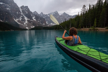 Adventurous woman kayaking in Moraine Lake during a striking cloudy sunset. Taken in Banff National...